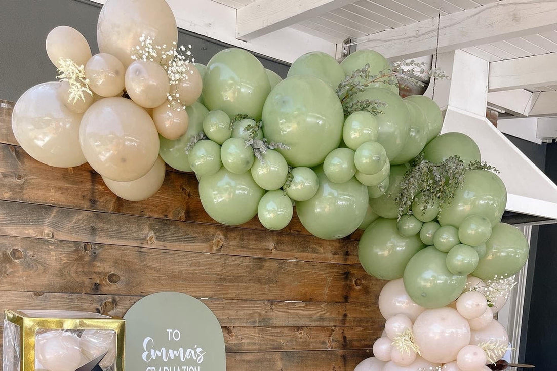 Sage green and cream balloon garland arching over a wood wall at a graduation party